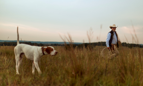 Man with cowboy hat on walking in a field with a hunting dog