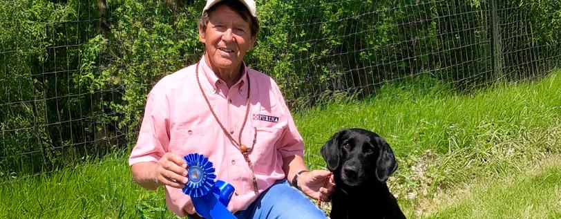 Bob West posing with medal and black lab in a field. 