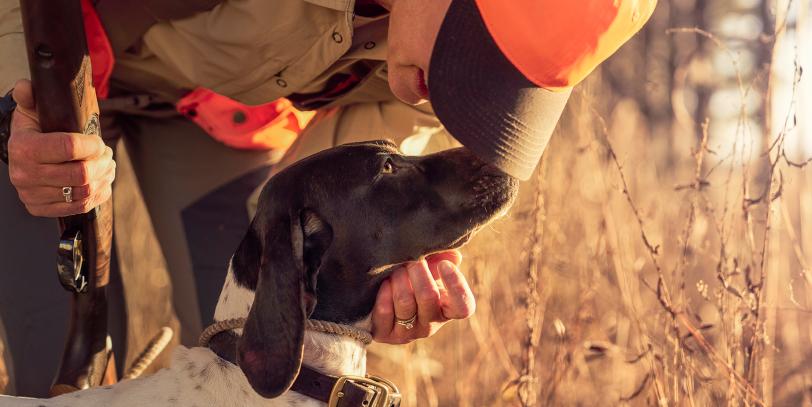A hunter holding a gun while bending down to talk to their dog.  